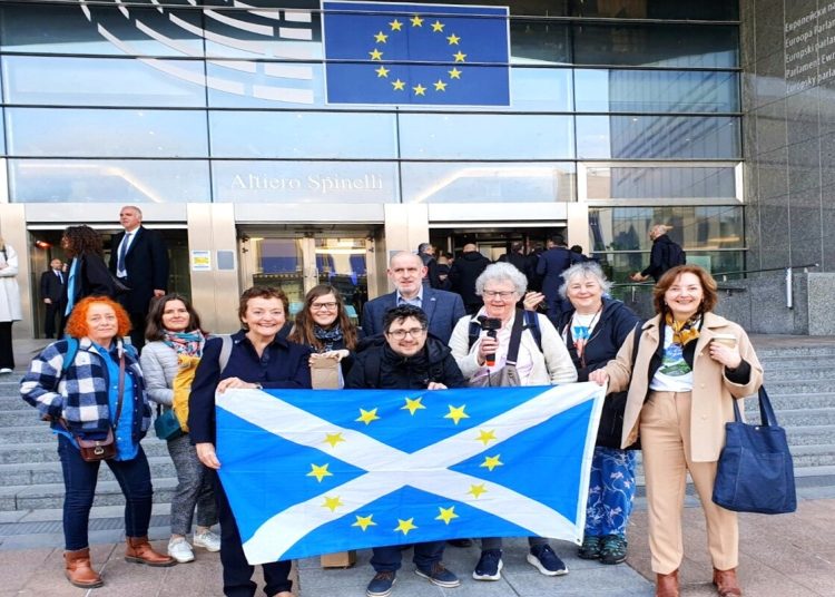 A photo of a group of pro-EU activists with Indy podcaster in front of the European Parliament bearing the Scottish flag with EU stars