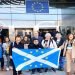 A photo of a group of pro-EU activists with Indy podcaster in front of the European Parliament bearing the Scottish flag with EU stars