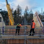 A photo of the MAKAR prototype house during construction. Two men in hi-vis vests and hard hats are standing on scaffolding and a crane can be seen in the background.
