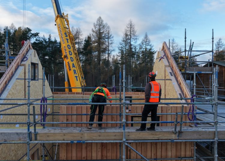 A photo of the MAKAR prototype house during construction. Two men in hi-vis vests and hard hats are standing on scaffolding and a crane can be seen in the background.