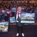 A picture of David Hayman, actor and activist, holding up signs with the words 'revive', 'what Scotland wants' and 'I back real land reform'. The photo was taken at Revive's National Conference 8 November 2025. In the background you can see the many visitors at the conference