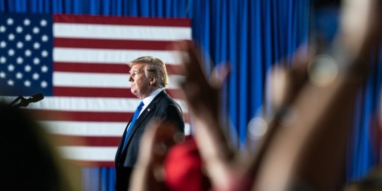 A photo of Donald Trump on stage with clapping hands in the front and the US flag against a blue curtain in the background.
