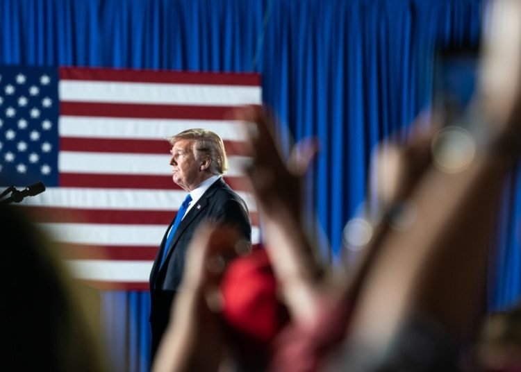 A photo of Donald Trump on stage with clapping hands in the front and the US flag against a blue curtain in the background.
