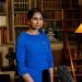 A portrait photo of Suella Braverman, then Home Secretary for the Conservative Party, in a library with wooden shelves and a switched on standard lamp to the right.
