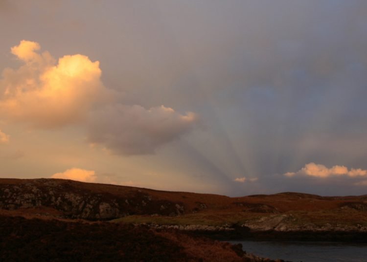 A photo of the sun setting over the Outer Herbrides casting shadows over the landscape. A cloud to the left is illuminated by the setting sun.