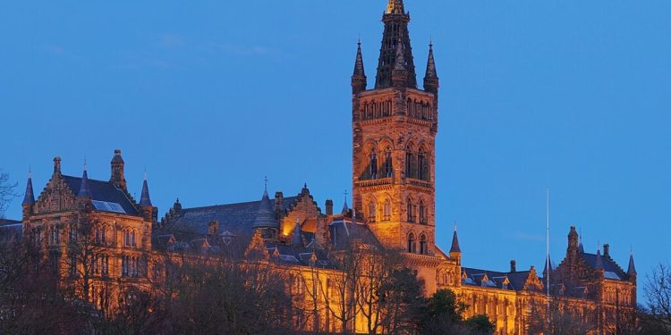 A photo of a the Gilbert Scott Building at the University of Glasgow floodlit from below against a dark blue evening sky.