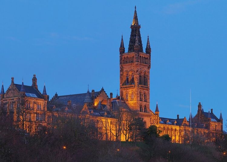 A photo of a the Gilbert Scott Building at the University of Glasgow floodlit from below against a dark blue evening sky.