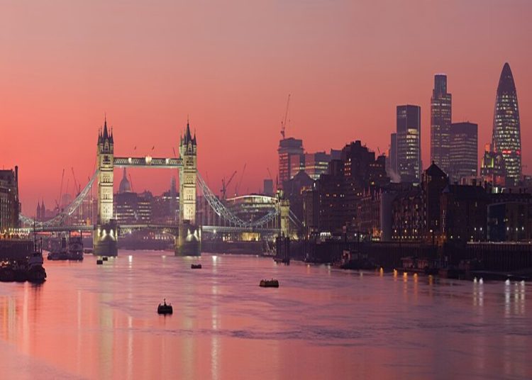 A photo of the city of London at sunset. The sky is a pink/orange and the river Thames is mirroring the colour of the sky. Tower Bridge is lit up and is to the left of the picture. CIty skyscrapers are to the right.
