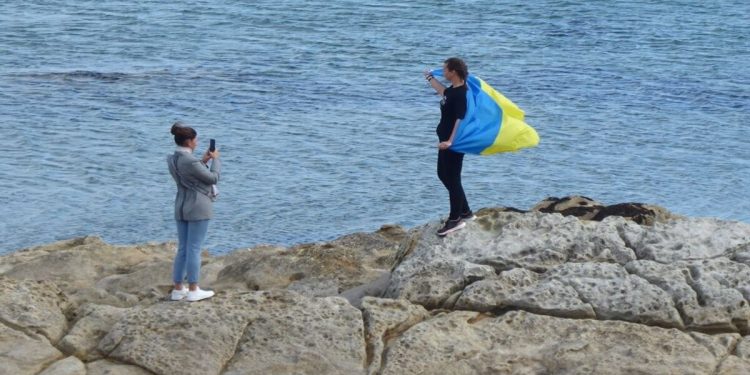 A photo of two people standing on a rocky part of the Scottish coastline with the sea in the background. The person on the left is taking a photo of the person on the right who has the Ukrainian flag draped over his shoulders.
