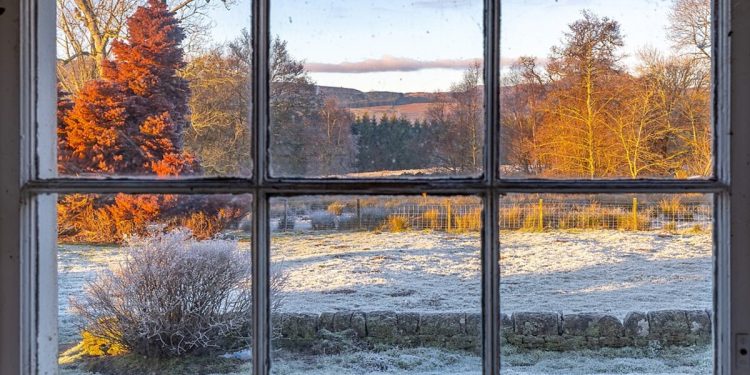 A photo looking through a wooden window into the frozen Scottish countryside. The wooden window is not in good repair and the glass panes are a little dirty. The sun is shining on the landscape outside lighting up the trees and perhaps symbolising a golden future where the frozen ground has thawed further away towards the horizon.