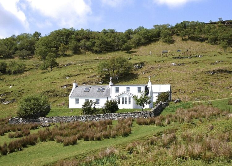 A photo of a farmhouse holiday letting property in Brochel, Raasay, Scotland. The white building is set on a hilly landscape with trees and shrubs dotting the ground. A light blue sky with white clouds can be seen at the top of the picture.