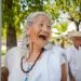 A photo of a grey-haired lady with a look of amused joyous surprise on her face. She is holding on to a pole and is wearing a white blouse and a wooden beaded necklace. In the background there is a grey-haired man with a hat looking at her. The image of him is slightly blurred.
