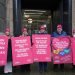 A photo of teachers holding protest posters reminding the Scottish Government of their workload and the need for recognition and love on Valentine's Day. All six protesters are wearing pink beanies matching the background of the placards they are holding in front of the Scottish Government building.