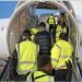 A photo of an asylum seekers being deported by the Home Office. The picture shows the back of a man in a black vest boarding the back steps of an aeroplane accompanied by six people in bright yellow hi-vis jackets.