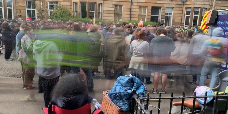 A photo of protesters at Kenmure Street in Glasgow. The middle part of the picture of the protesters is blurred. In the background the iconic stone tenement buildings typical for Glasgow.