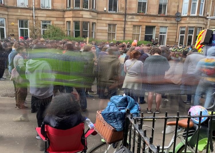 A photo of protesters at Kenmure Street in Glasgow. The middle part of the picture of the protesters is blurred. In the background the iconic stone tenement buildings typical for Glasgow.