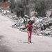 A girl dressed in red walks through the streets of Gaza looking for food, she is holding a pot in her right hand, she is surrounded by rumble from destroyed buildings