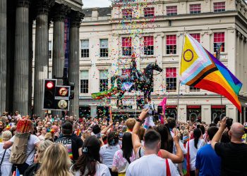 Crowd at a Pride parade with a large rainbow flag, confetti raining down, and a statue of a horse in the background.
