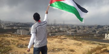 Man on a hill with his back facing the reader and waving a palestinian flag
